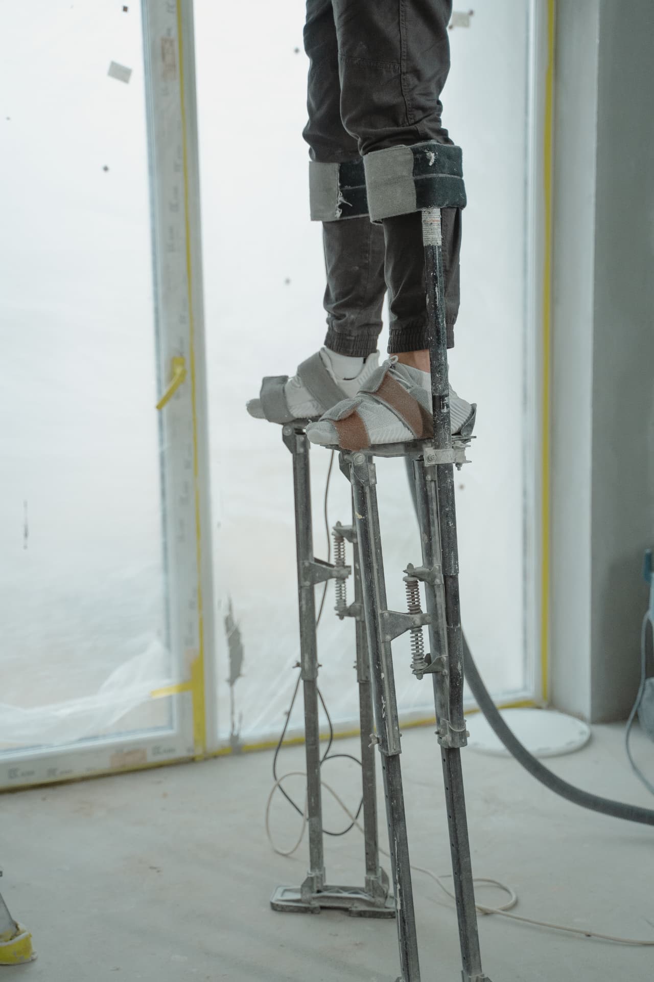 A contractor standing on aluminum drywall stilts inside an unfinished room