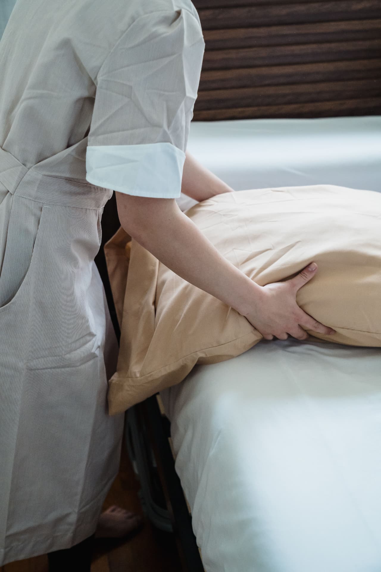 A housekeeper in a tan uniform placing a beige pillow on a bed with crisp white linens